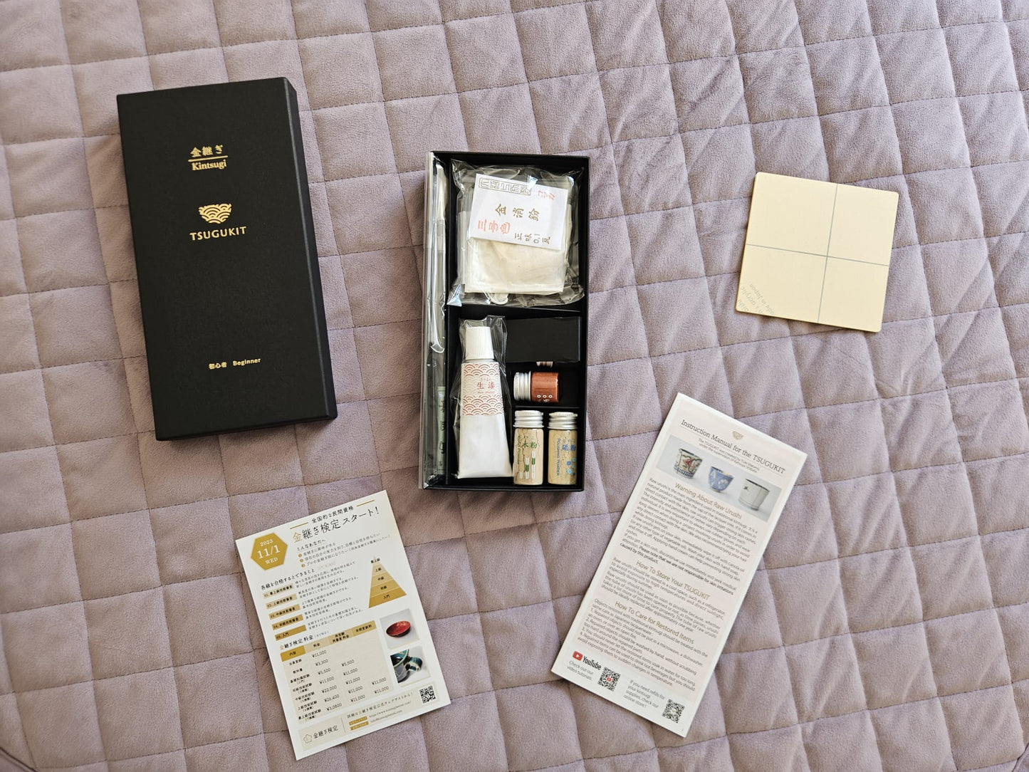 Kitntsugi kit set for beginners featuring a black box, instructions, ink, palette dropper, spatula, gold powder, masking tape on a purple background.